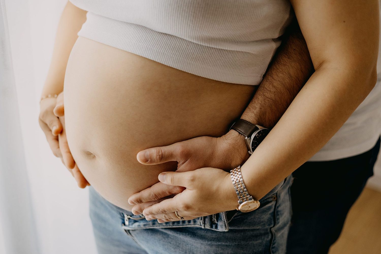 Close-up of pregnant belly with couple's hands cradling bump, wearing jeans and watches.