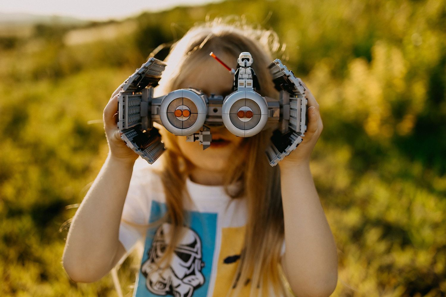 Young girl holding a LEGO spaceship toy up to her face like binoculars in a green field.