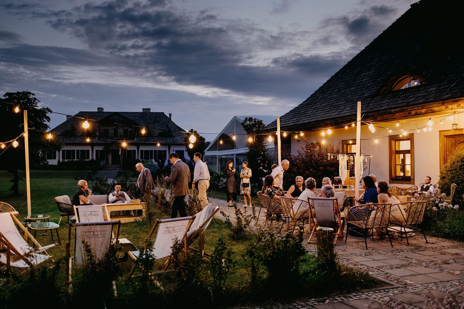 Outdoor wedding reception at dusk with string lights and guests socializing.