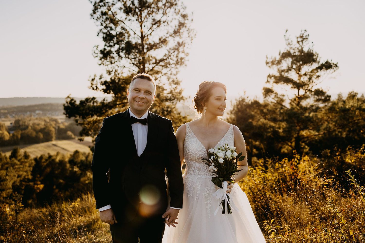 Smiling bride and groom holding hands at golden hour with a sunlit forest landscape behind them.