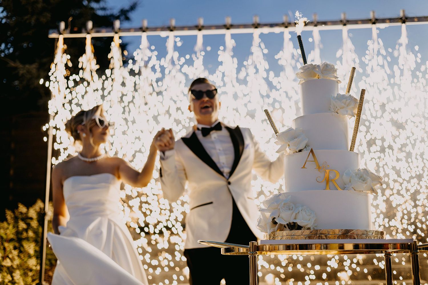 Bride and groom celebrating by a tiered white wedding cake with gold details and sparkling backdrop.