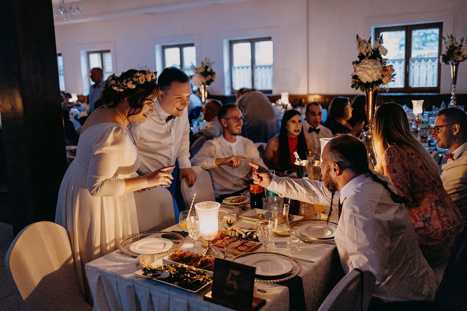 Bride and groom interact warmly with seated guests at candlelit wedding reception dinner in a rustic venue.