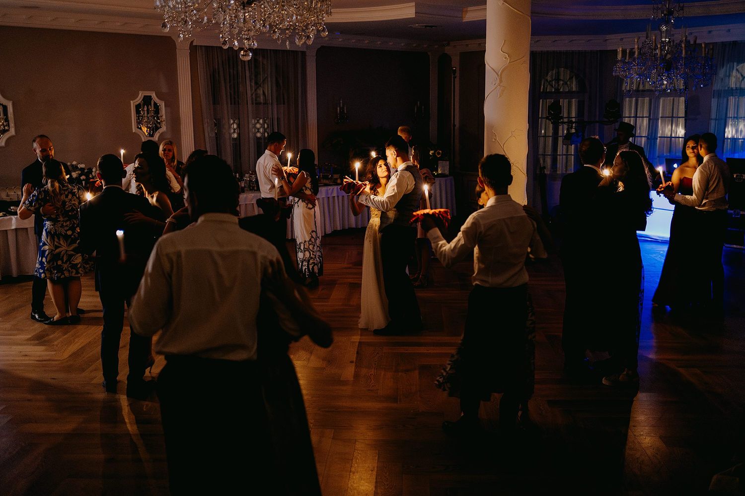 Guests dancing by candlelight in an elegant dimly lit wedding reception hall with chandeliers.