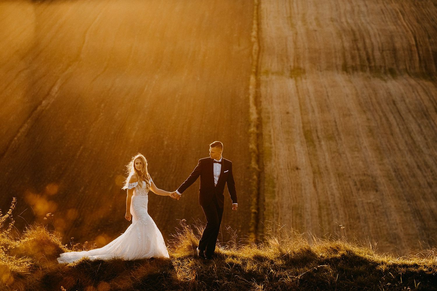 A bride and groom walk hand in hand through a sunlit golden field at sunset.