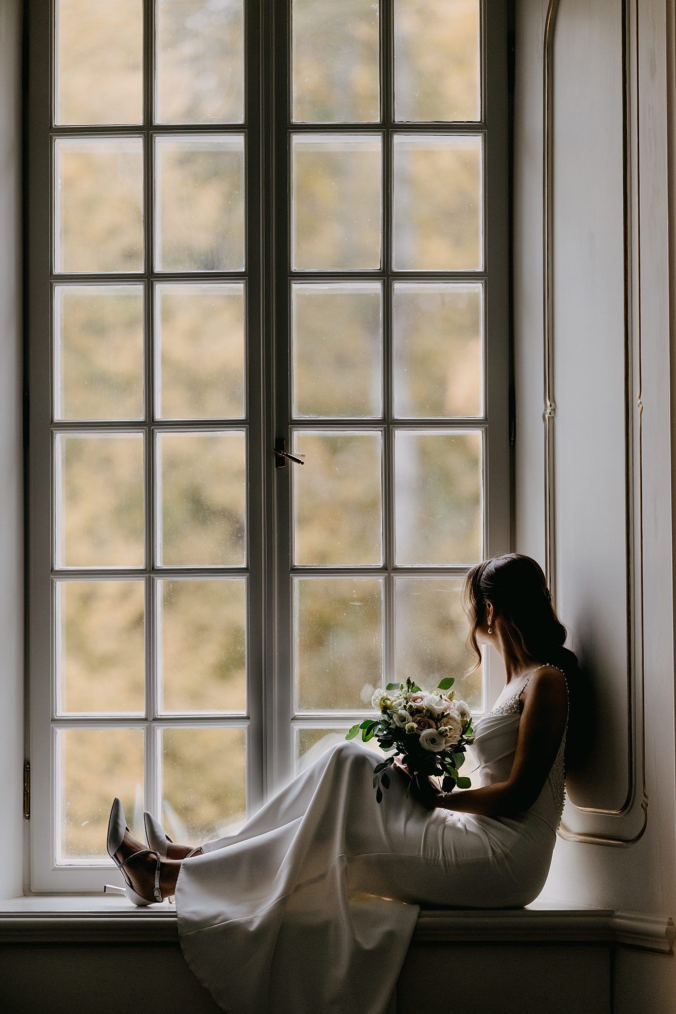 Bride in white gown sitting on a windowsill holding a bouquet, gazing out at autumn foliage.