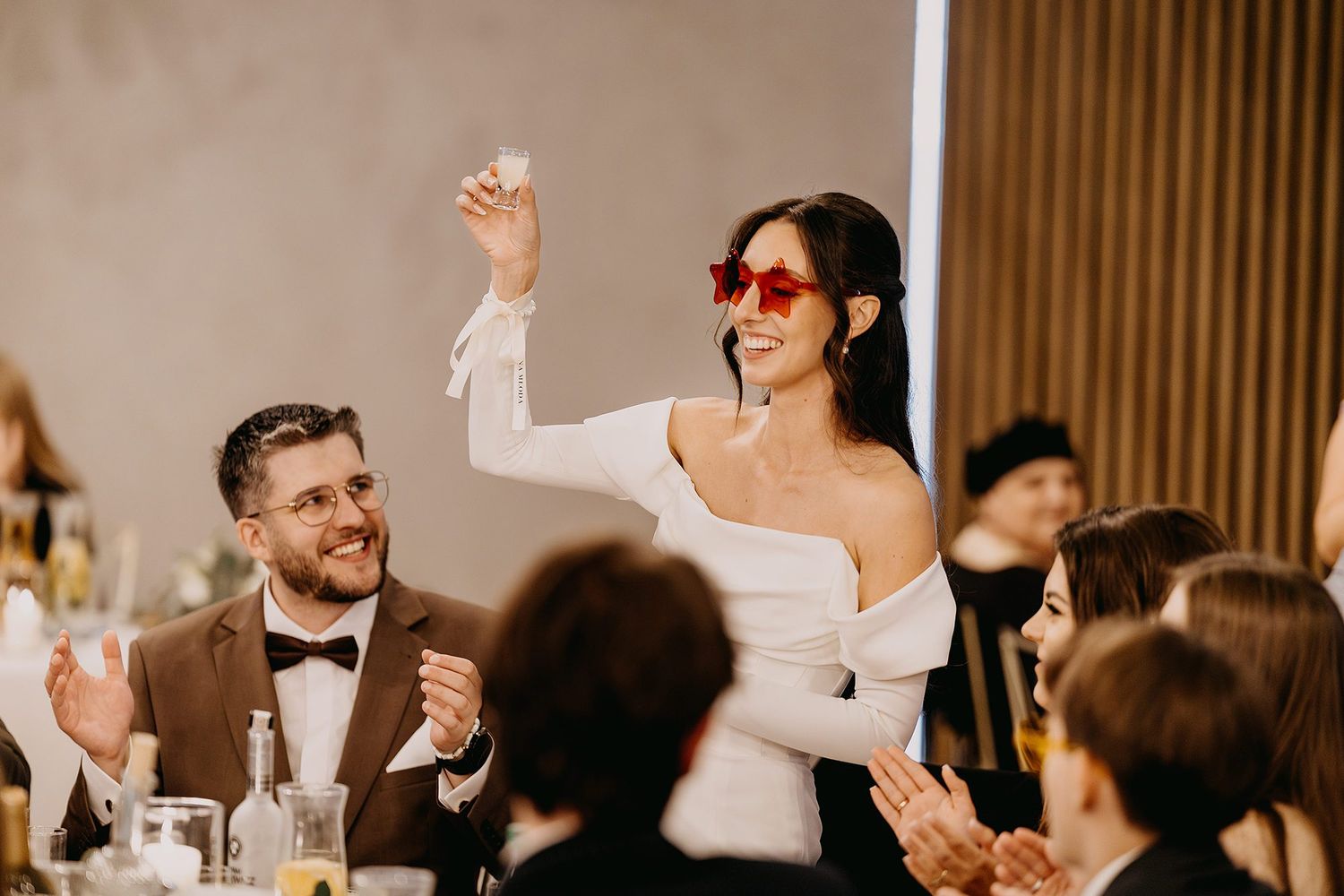 Bride in white off-shoulder gown wearing red sunglasses raises glass while guests applaud at reception.