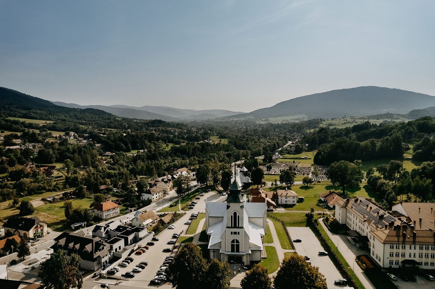 Aerial view of a small European town with a white church surrounded by green hills and mountains.