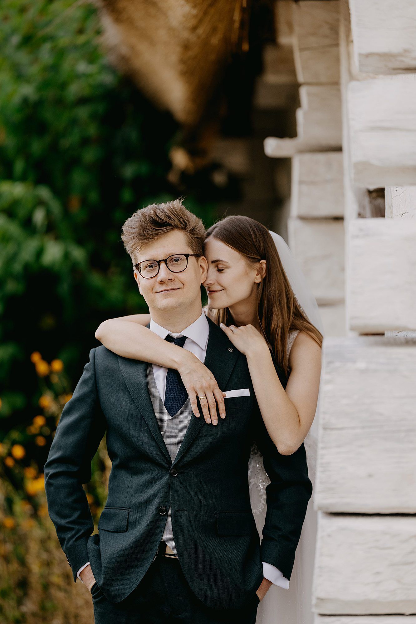 Bride embracing groom in glasses from behind near a rustic stone building with greenery.