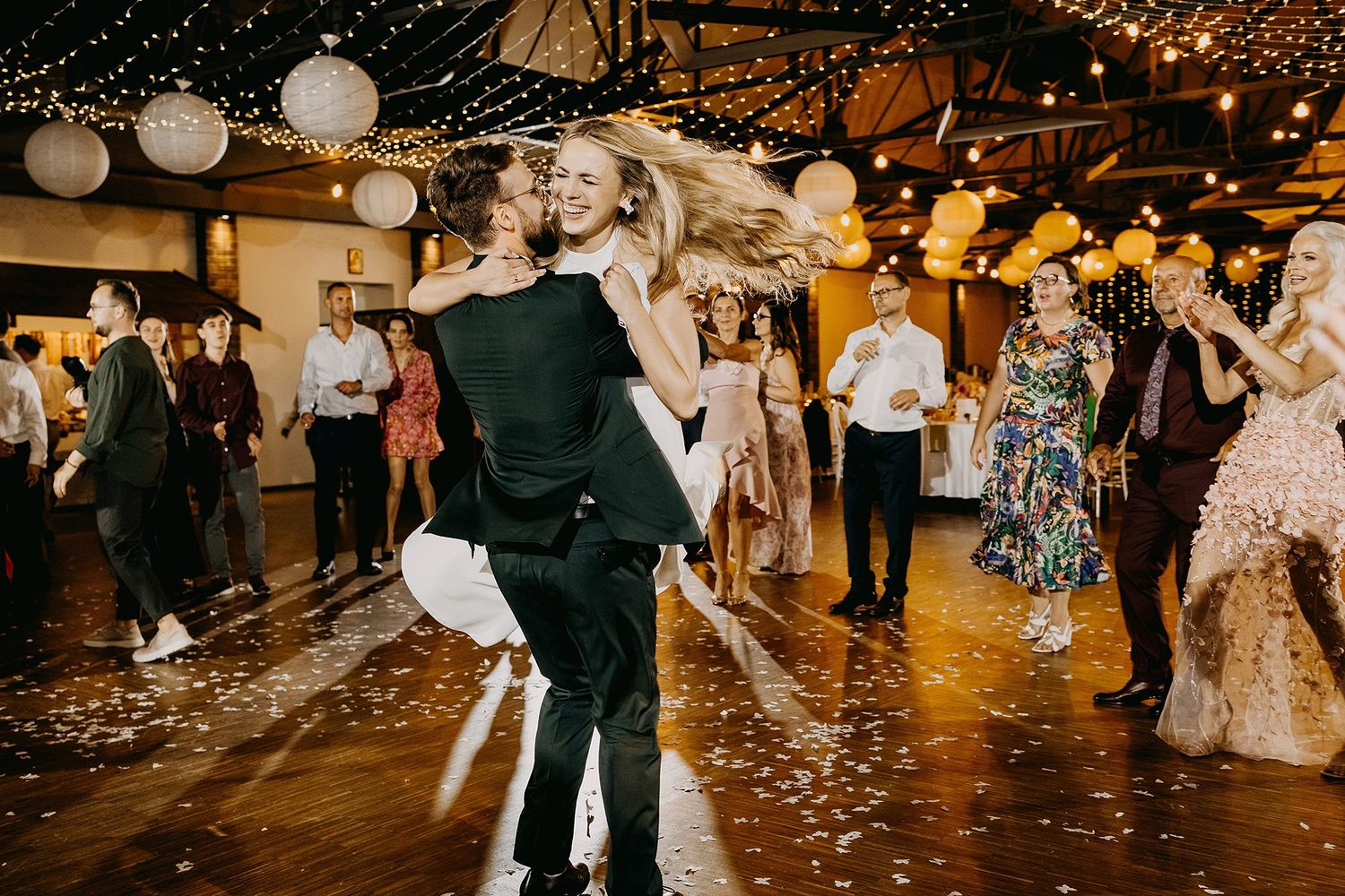 Couple sharing joyful first dance at wedding reception with guests, lanterns, and gold balloons in background.