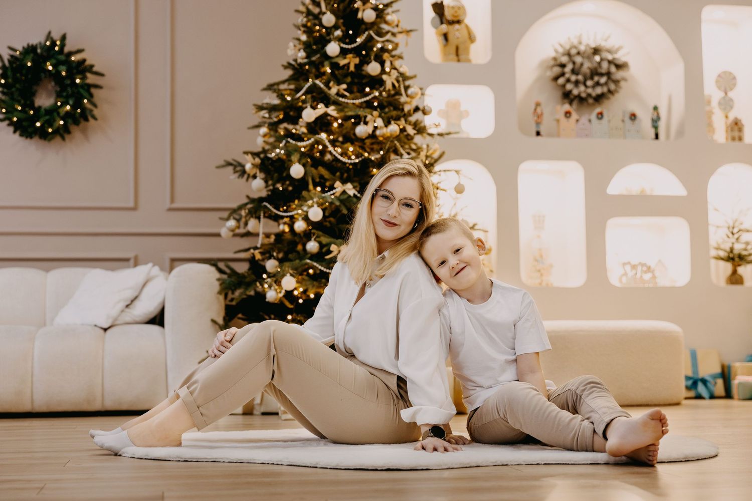 Mother and young son in cream outfits sit together on a rug beside a decorated Christmas tree in a bright, cozy living room.