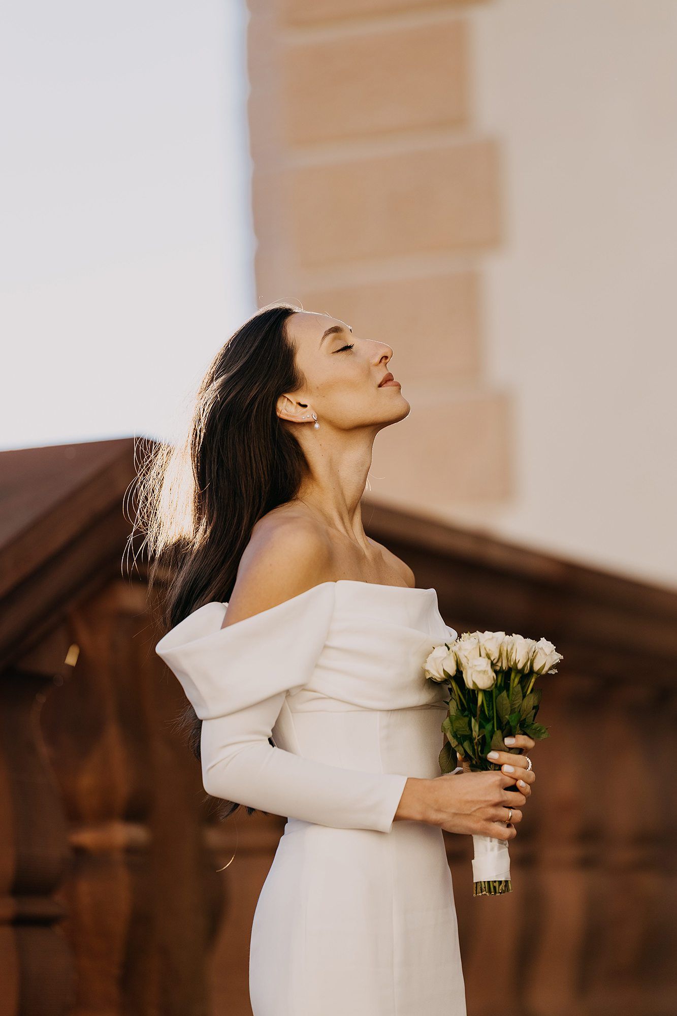Elegant bride in off-shoulder white gown holds white tulip bouquet with eyes closed peacefully.