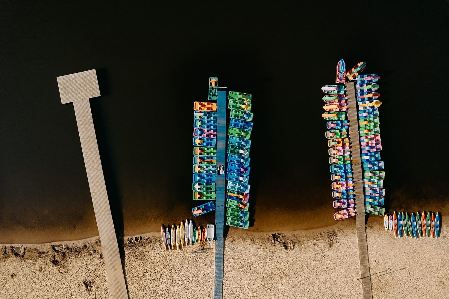 Aerial drone view of colorful paddleboats and kayaks docked in rows along a dark sandy shoreline.