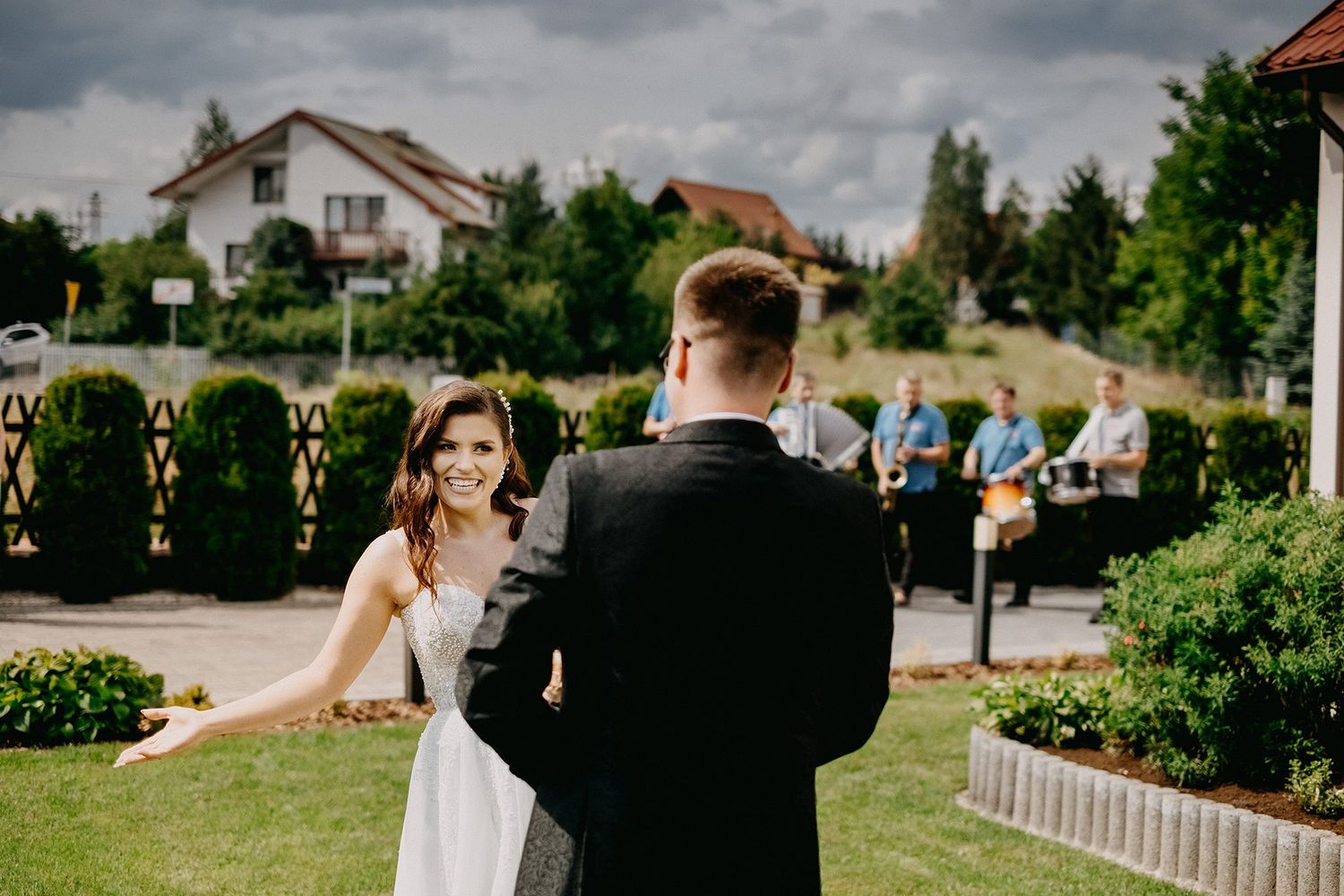 Smiling bride in white gown spreading arms to greet groom outdoors at a garden wedding venue.