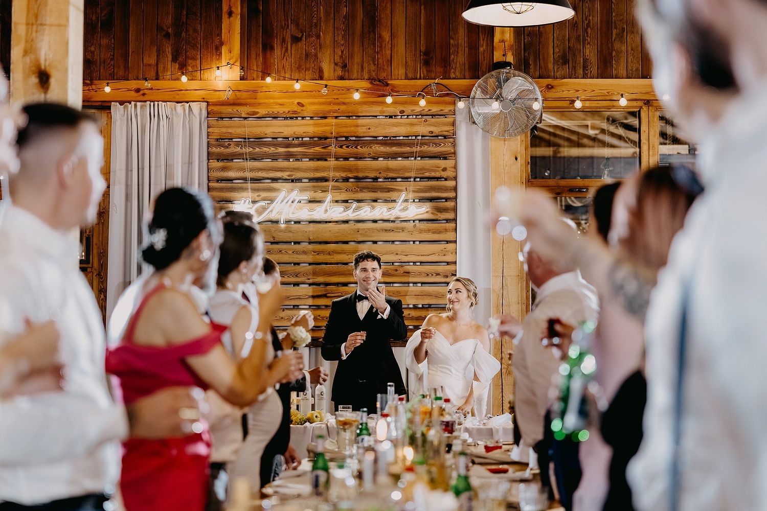 Bride and groom share toast with guests in rustic wooden barn reception hall.