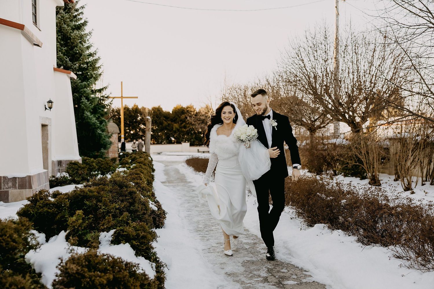 Happy newlyweds walk together along a snowy path outside a church in winter wedding attire.