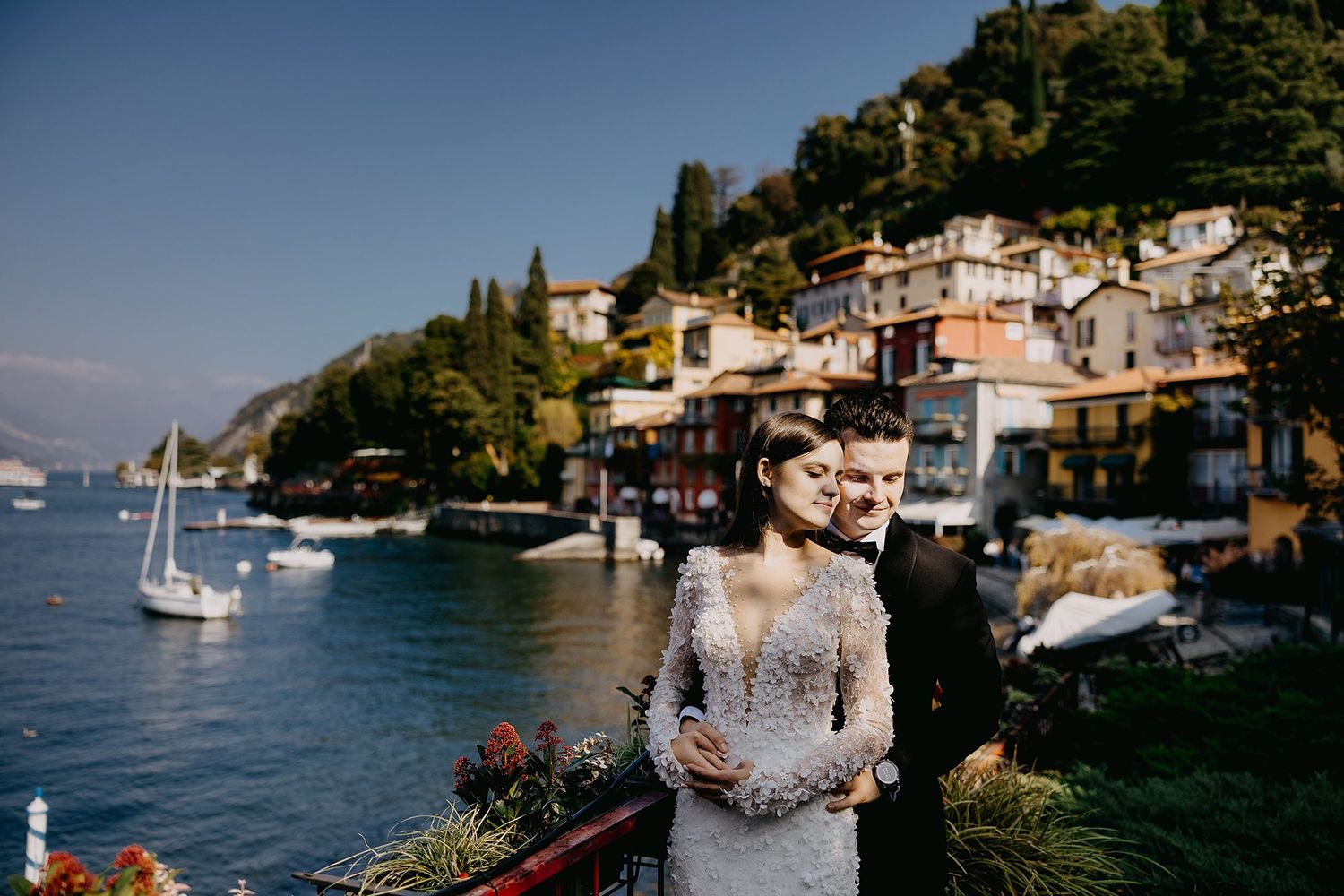 Bride and groom embrace on terrace with stunning Lake Como village and mountains in the background.