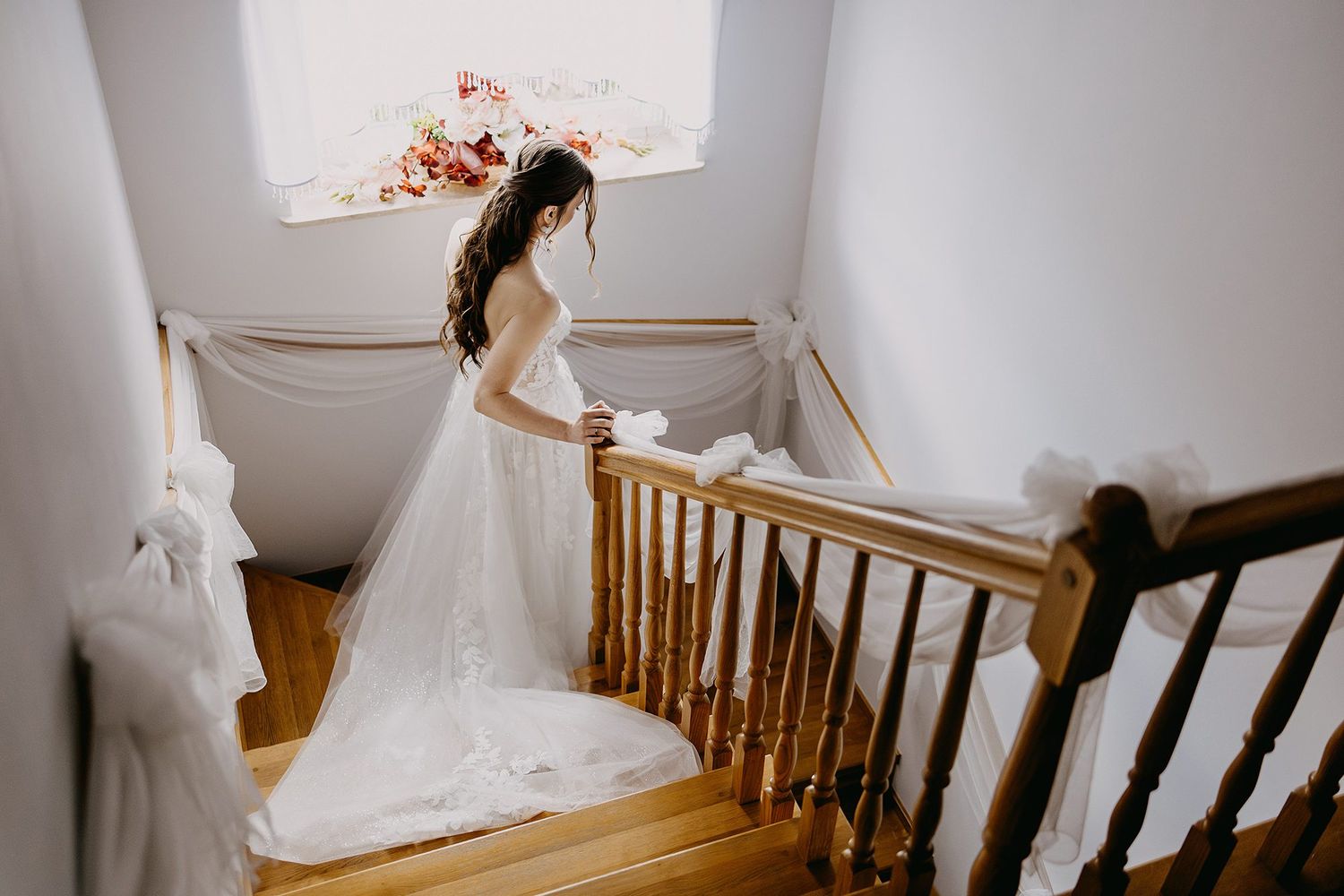 Bride in a lace wedding gown descending a wooden staircase with flowing train.