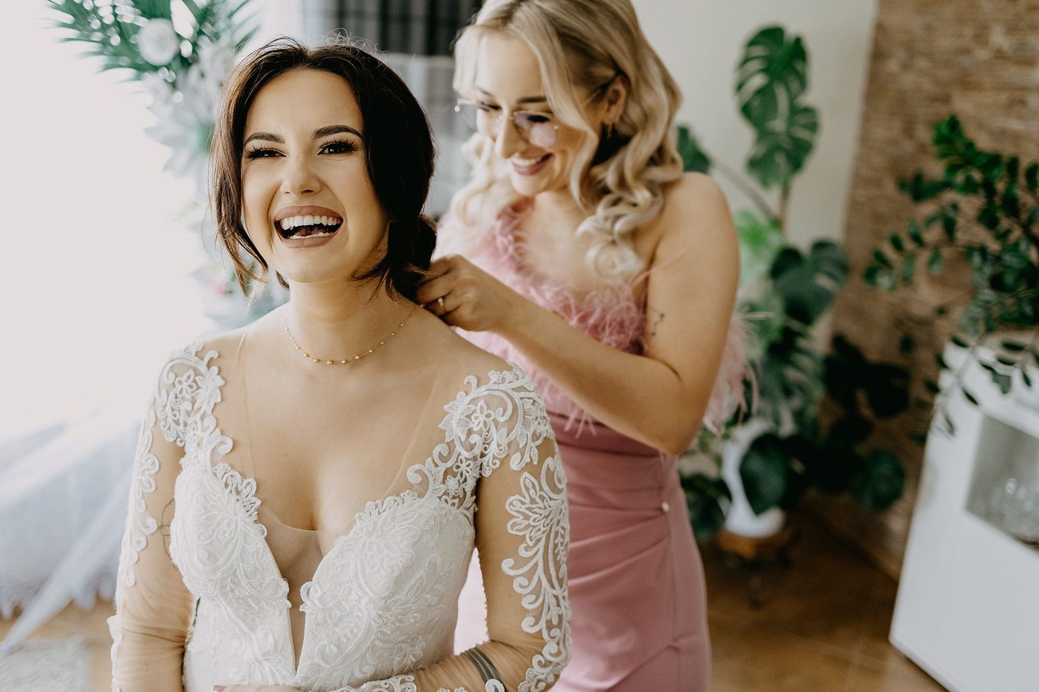 Bridesmaid in pink dress helping bride with necklace while both laugh joyfully.