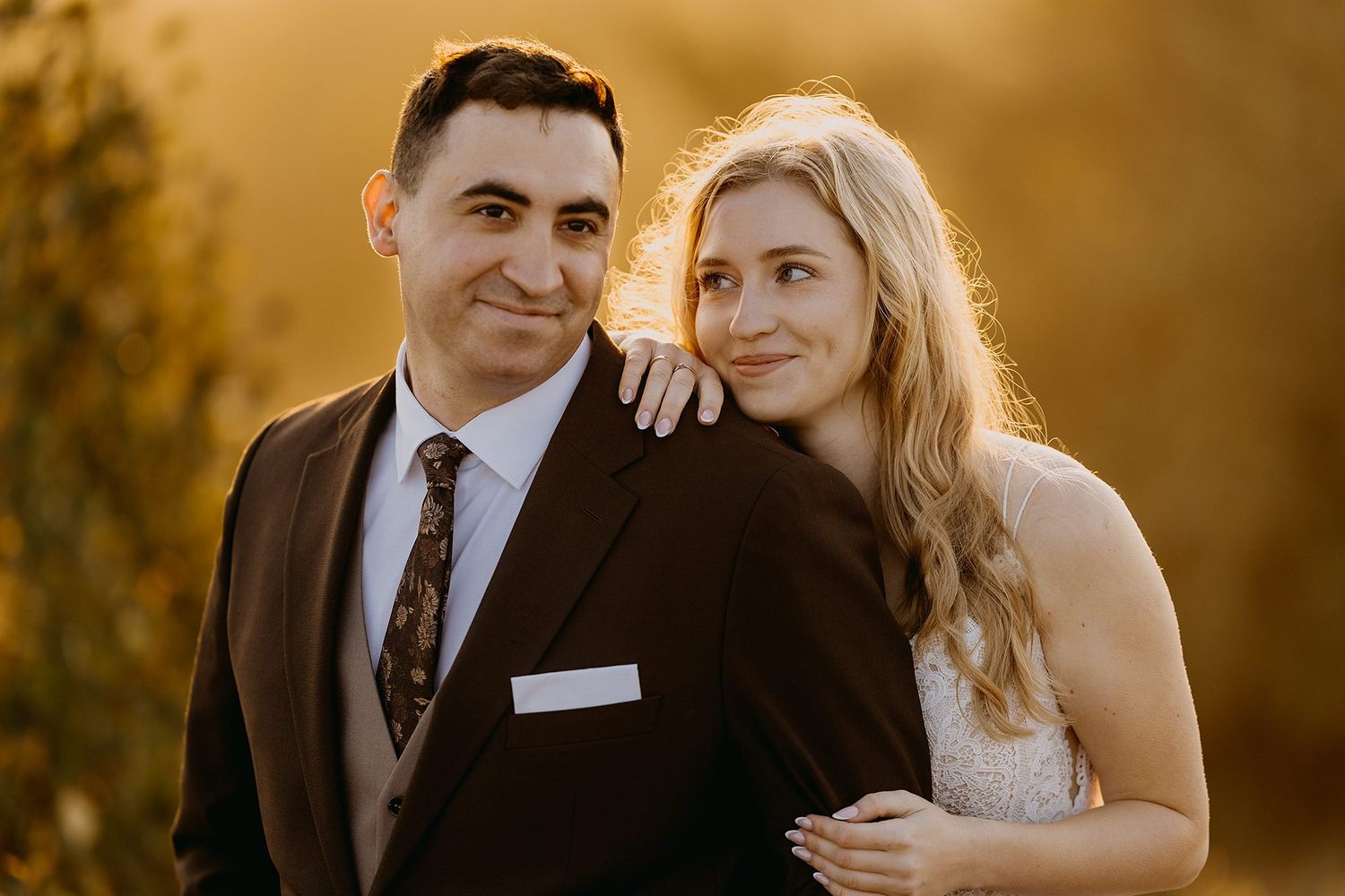 Bride leaning on grooms shoulder in a warm golden sunset outdoor wedding portrait.