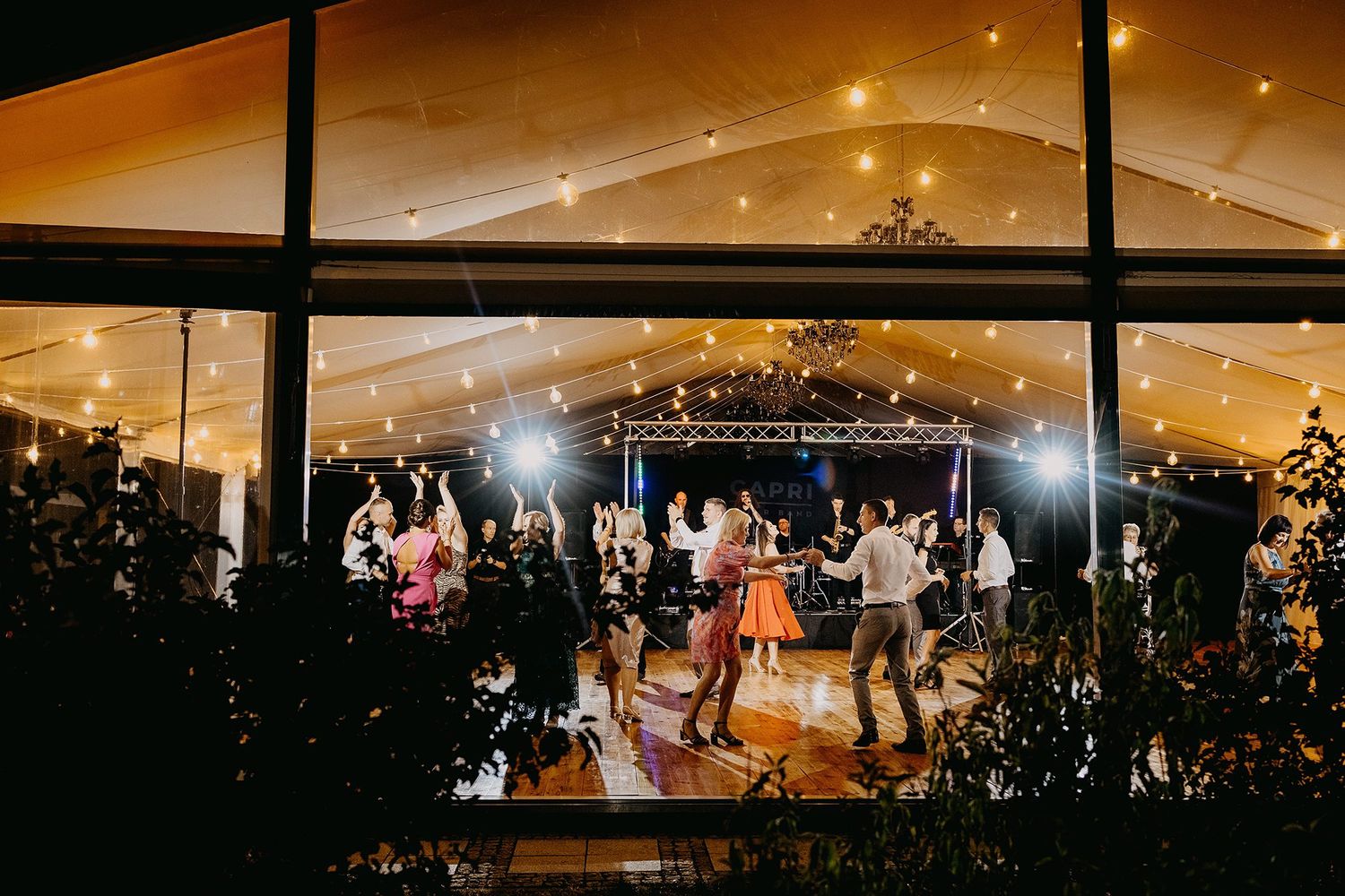 Guests dancing under fairy lights inside an elegant glass-walled wedding reception tent at night.