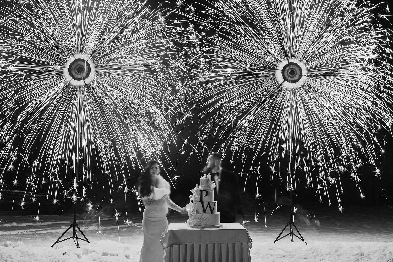 A couple cuts their wedding cake as spectacular fireworks explode dramatically behind them in black and white.