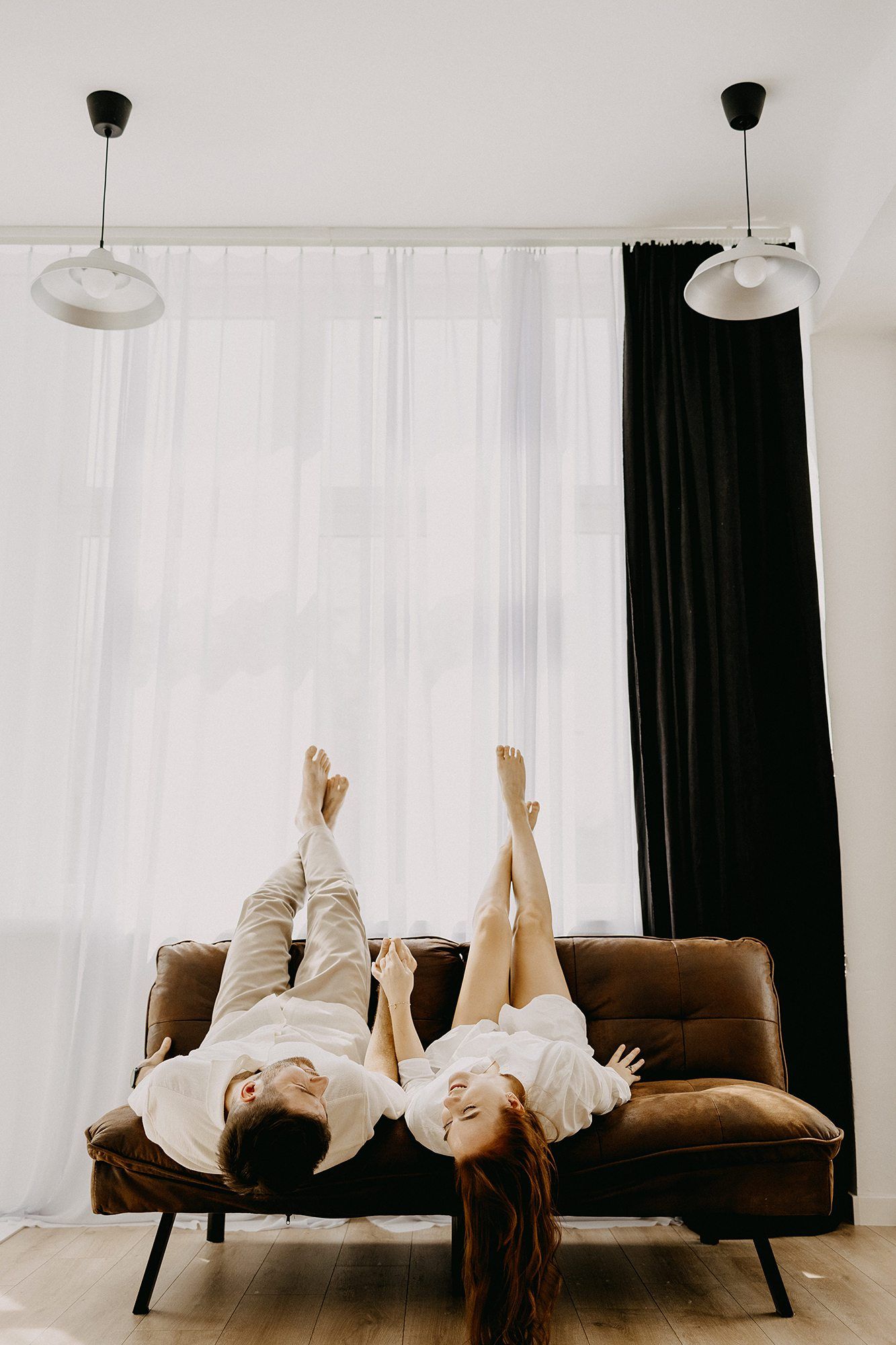Couple lying upside down on brown sofa near bright window.