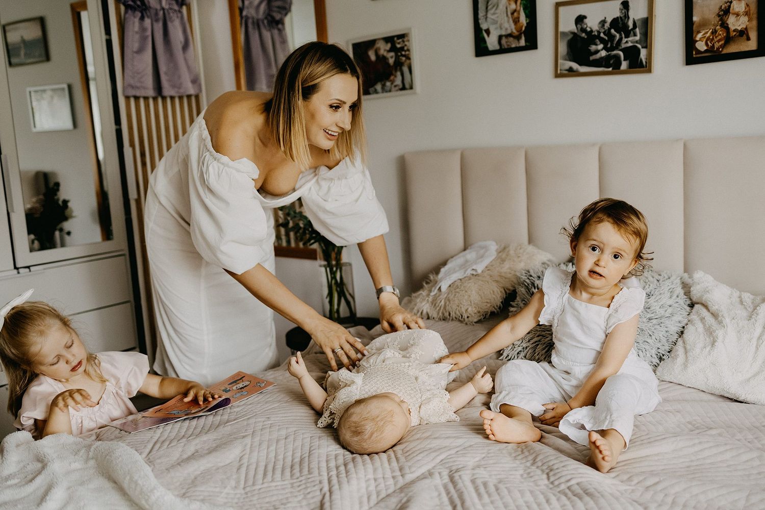Young mother in white dress plays with two toddler girls on a cozy bed in a bright bedroom.