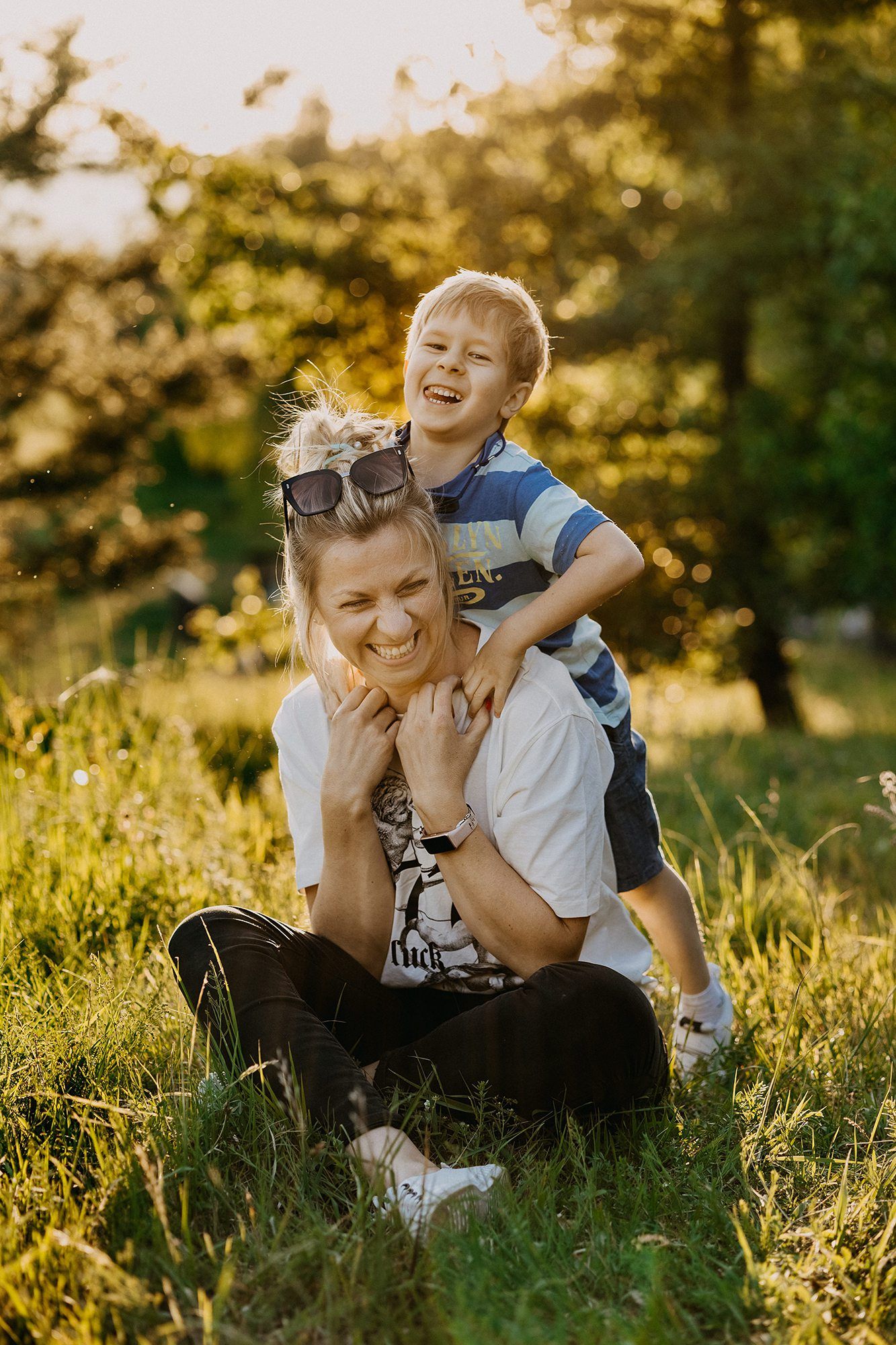 Laughing boy climbs on smiling mothers shoulders while sitting on sunny green grass.