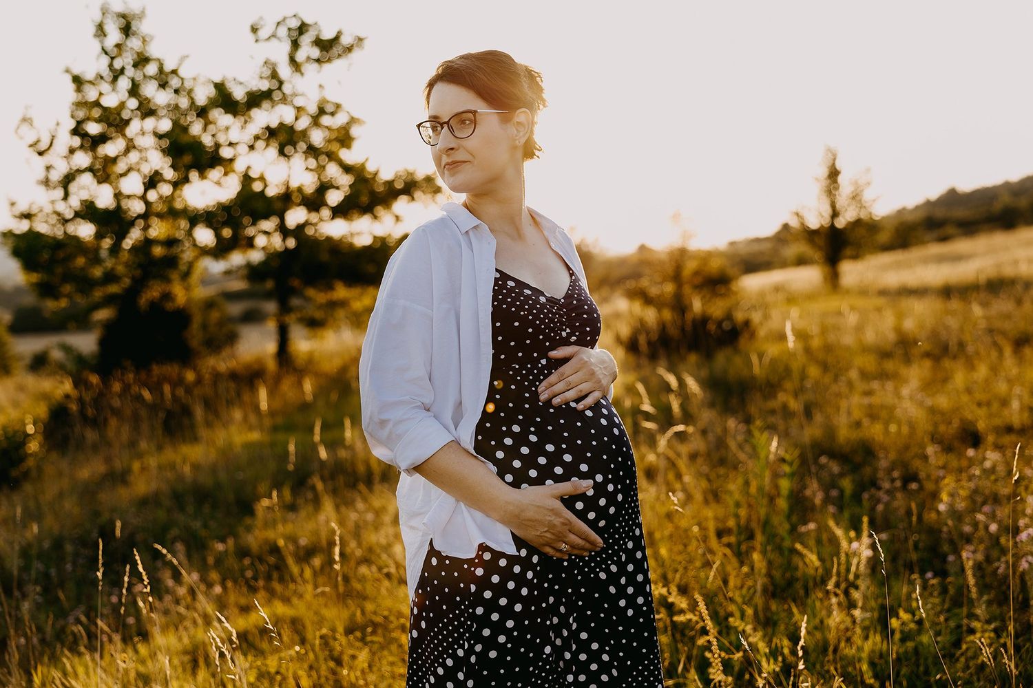 Pregnant woman in polka dot dress and glasses holding baby bump in sunny countryside meadow.