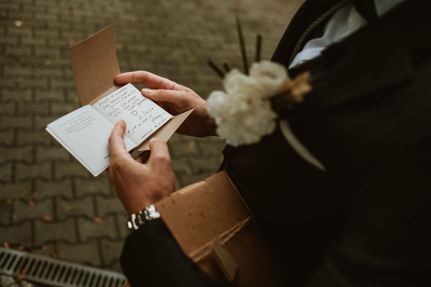 Groom in suit reading handwritten wedding note while holding kraft paper gift with white floral boutonniere.