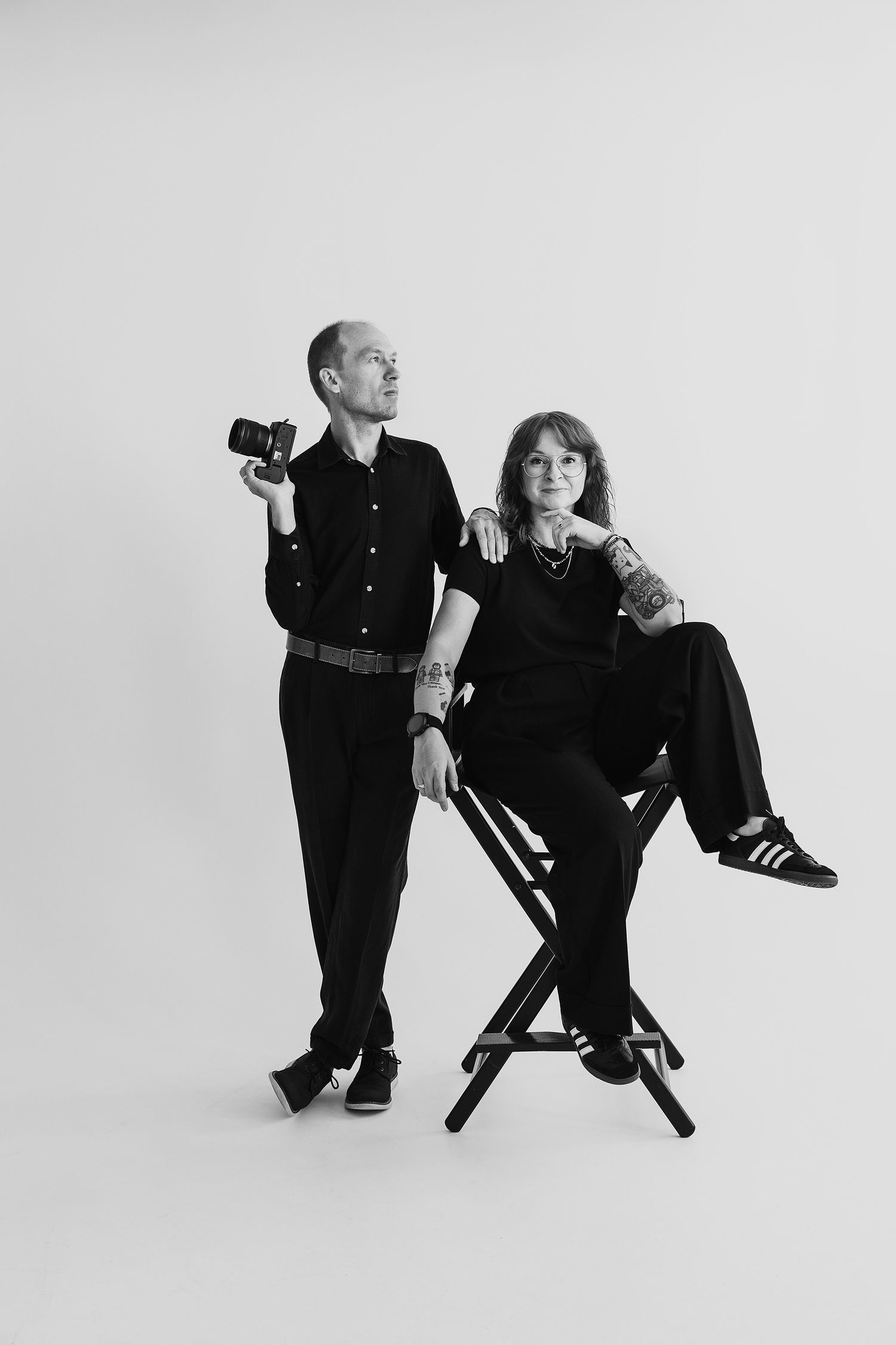 Black and white photo of two photographers in black outfits, man holding camera standing beside woman seated on director's chair.