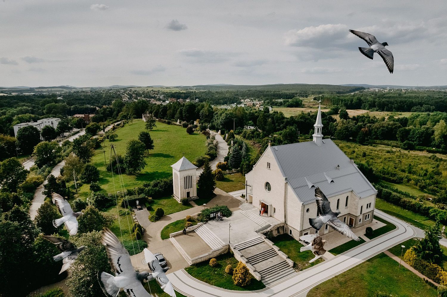 Aerial view of a historic church and chapel surrounded by lush greenery, with two birds flying overhead.