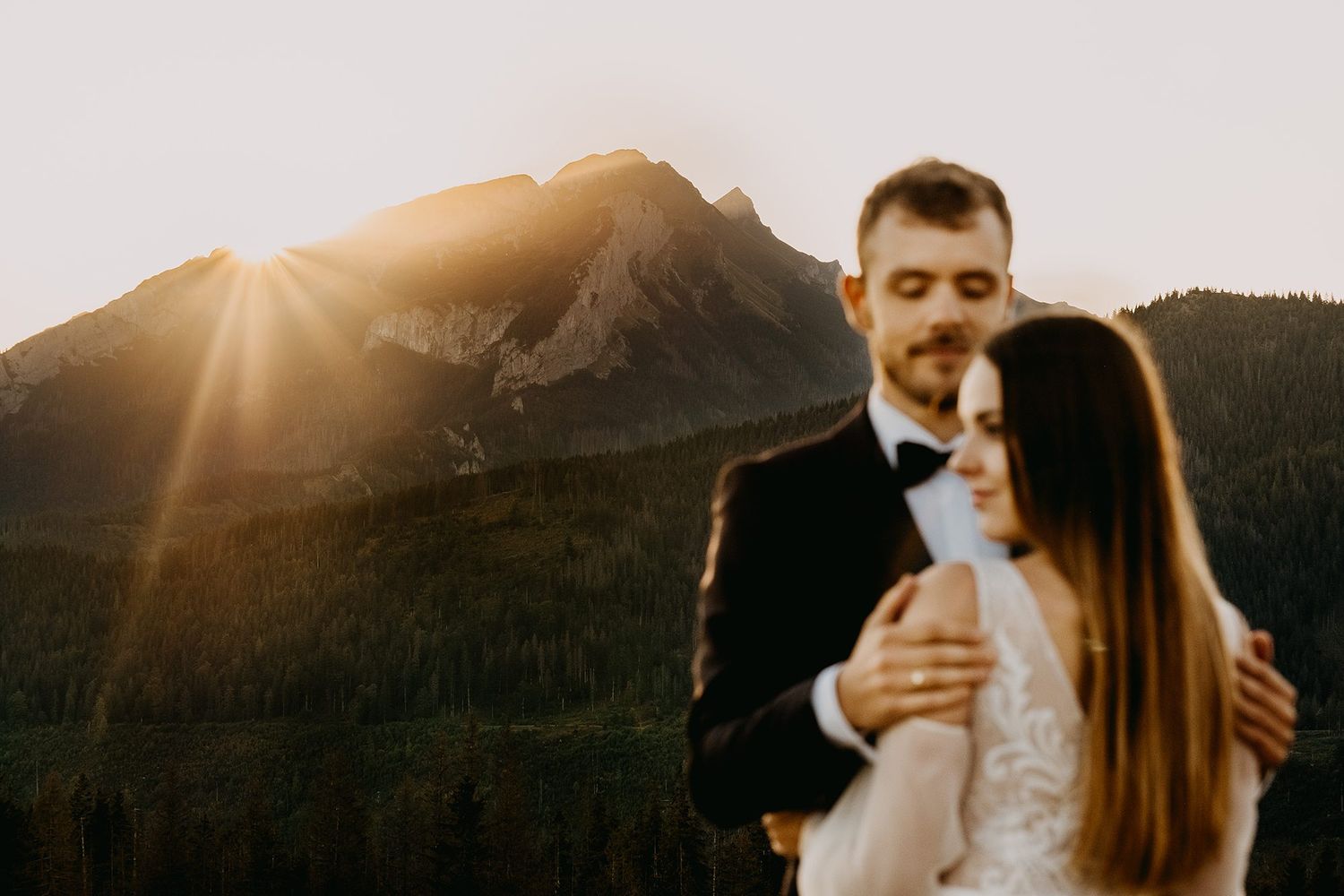 Groom tenderly embracing bride from behind with dramatic mountain peak and golden sunset behind them.