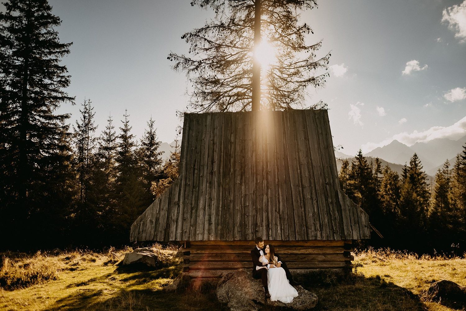 Couple embraces beside a rustic wooden chapel with sunlight streaming through tall pine trees.