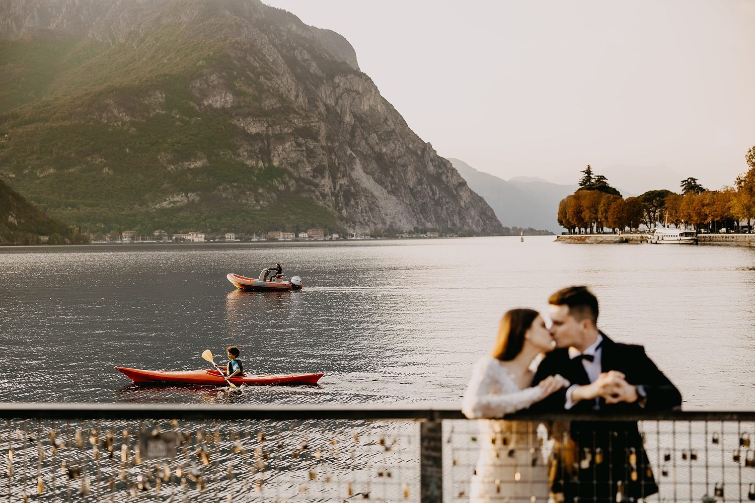 Bride and groom share a kiss on a lakeside promenade with kayakers and mountains behind them.