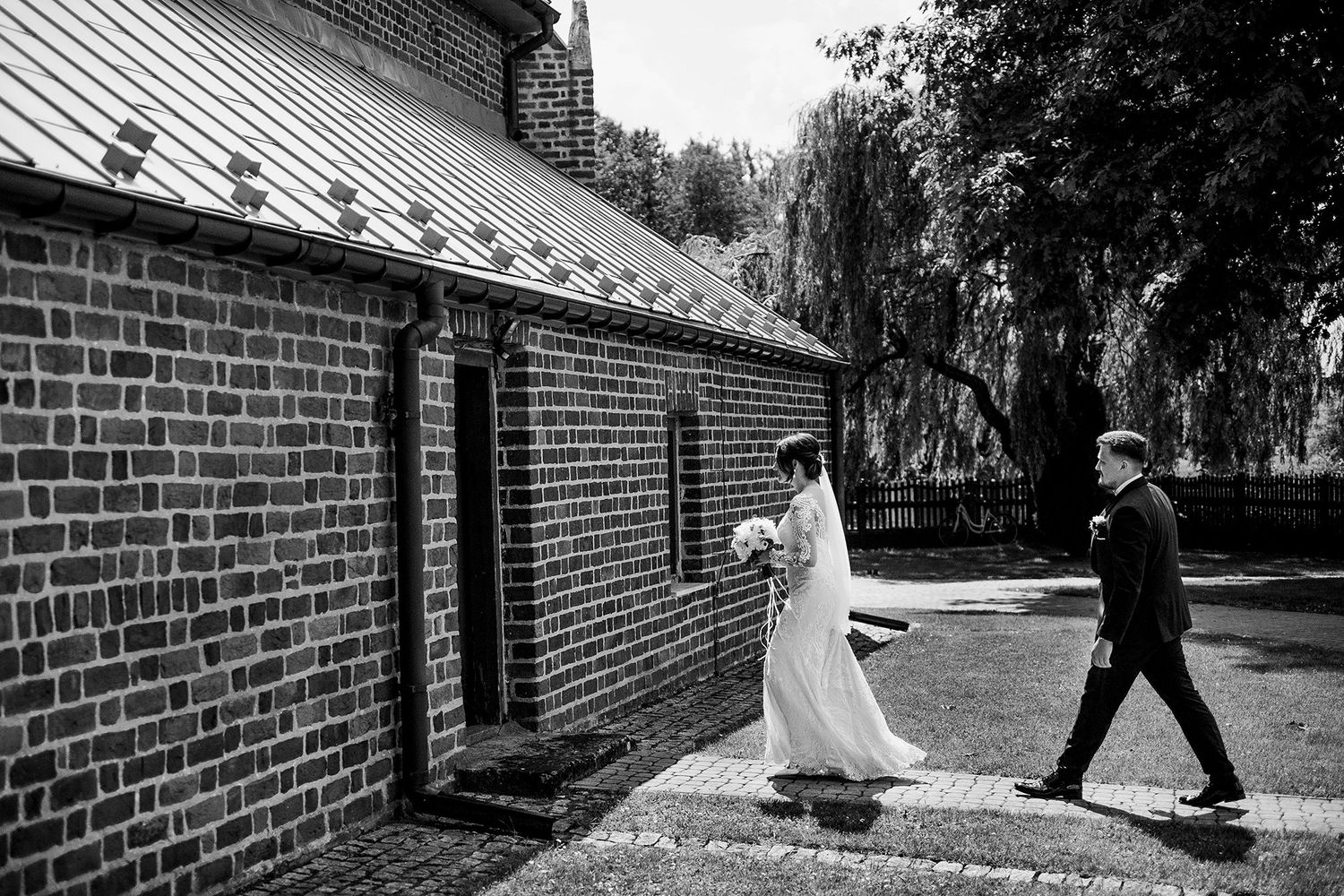 Black and white photo of bride and groom walking beside a brick church.