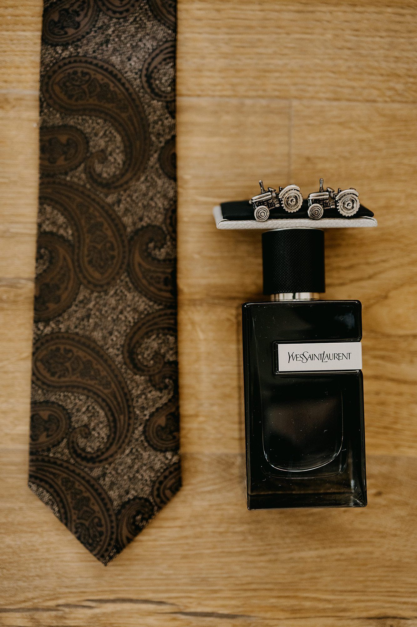 Yves Saint Laurent cologne bottle with tractor cufflinks beside a dark paisley tie on a wooden surface.