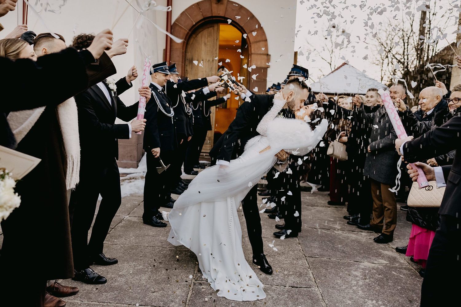 Bride and groom share romantic dip kiss outside church as guests shower them with confetti in winter.