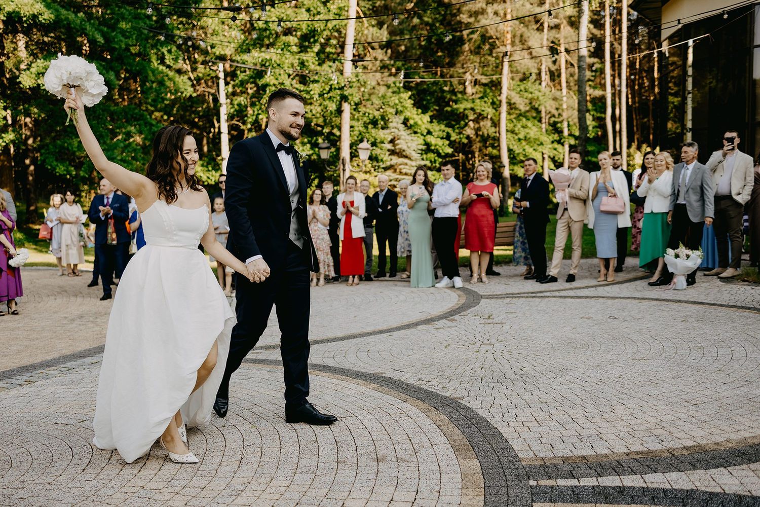 Joyful bride raising bouquet with groom on decorative paved outdoor wedding venue.