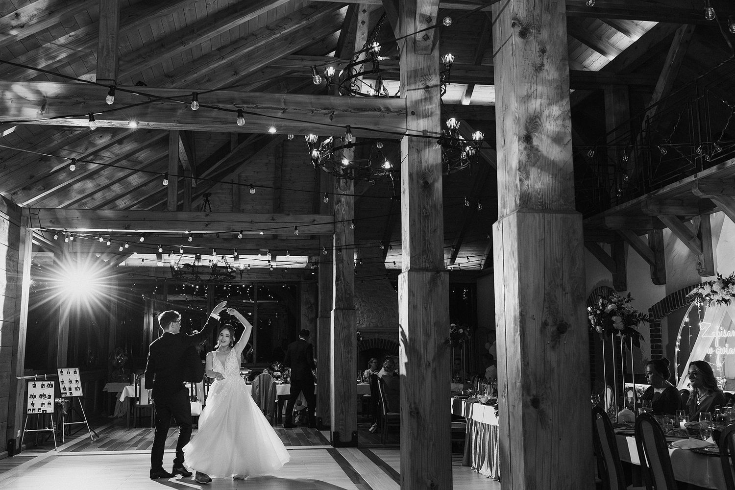 Black and white photo of bride and groom sharing first dance in rustic barn wedding venue with string lights.