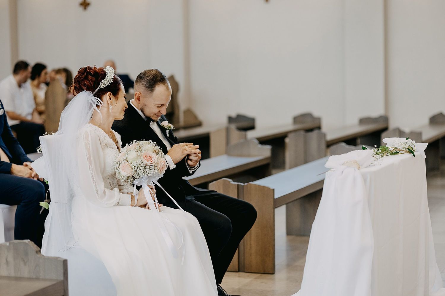 Bride and groom sitting together on church pew during wedding ceremony with floral decorations.