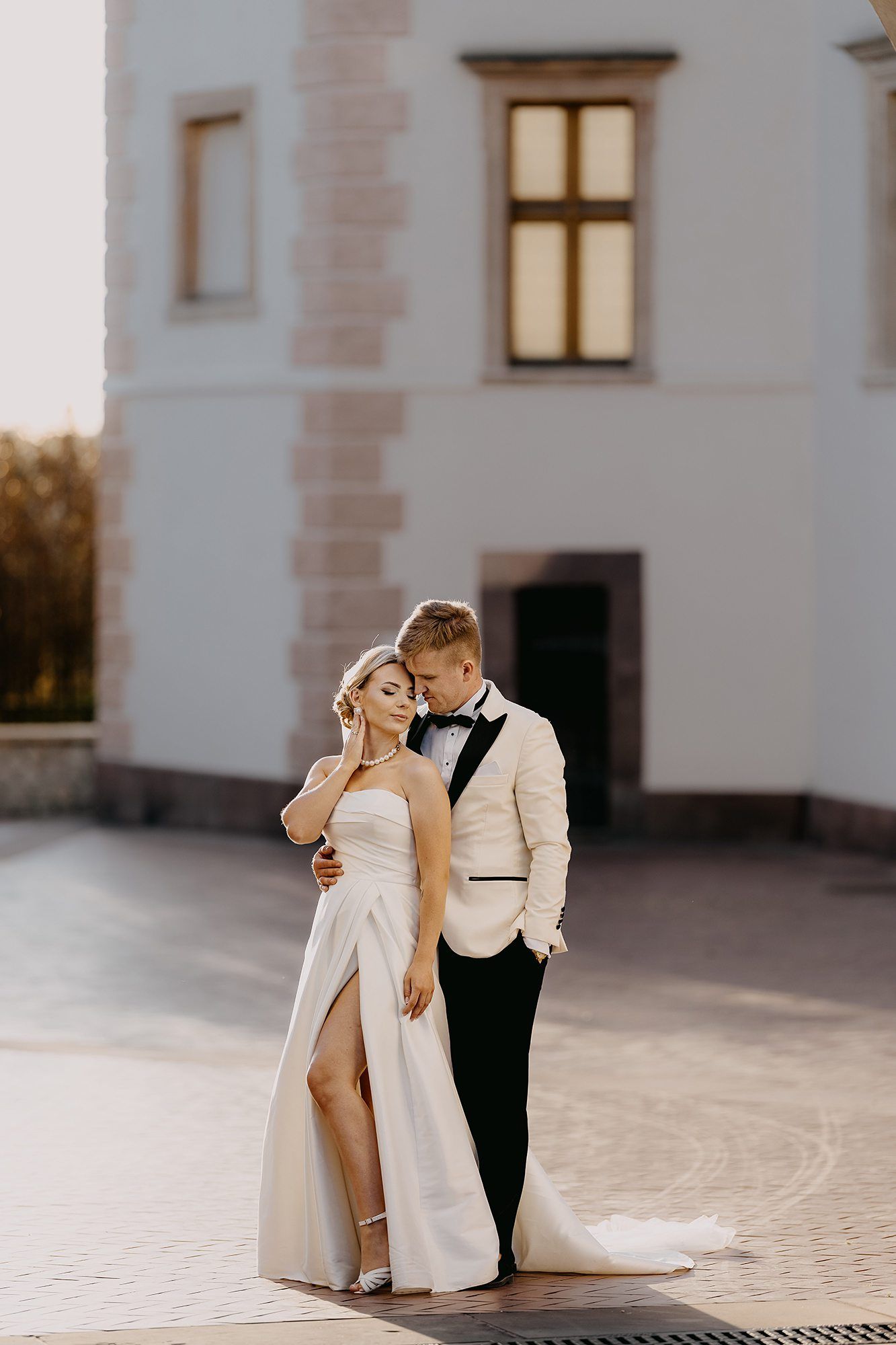 Groom in white tuxedo embracing bride in strapless gown outside an elegant white building.