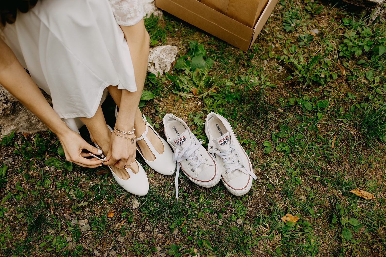 Bride sitting on grass choosing between elegant heels and white sneakers outdoors.