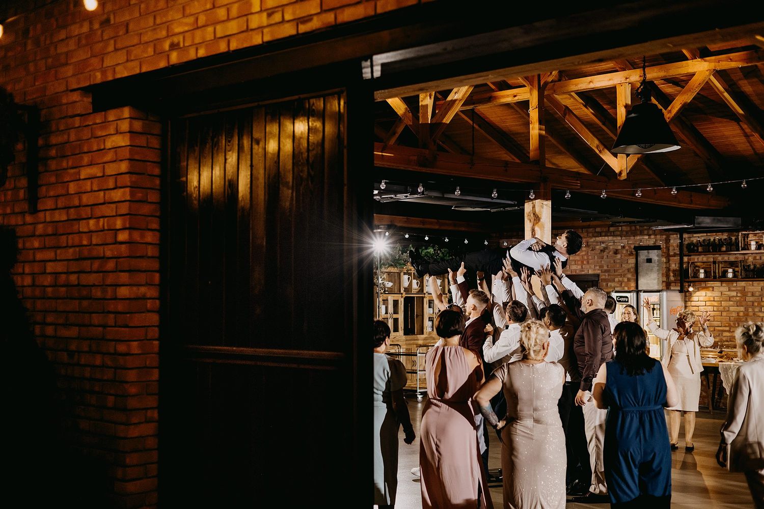 Wedding guests gathered at the entrance of a warmly lit rustic brick venue with exposed wooden beams.