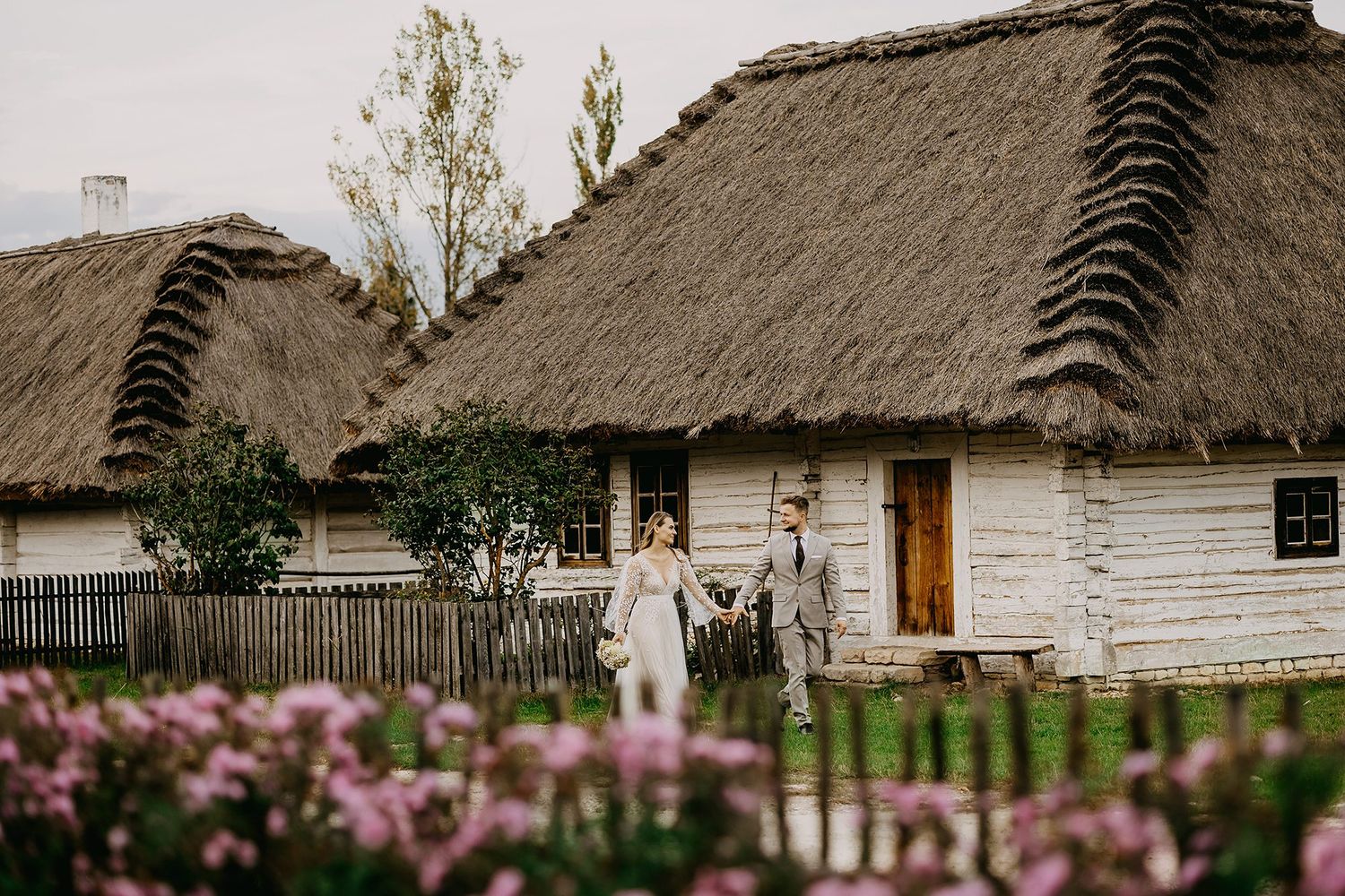 Wedding couple walks hand in hand past charming thatched-roof cottages surrounded by pink flowers.
