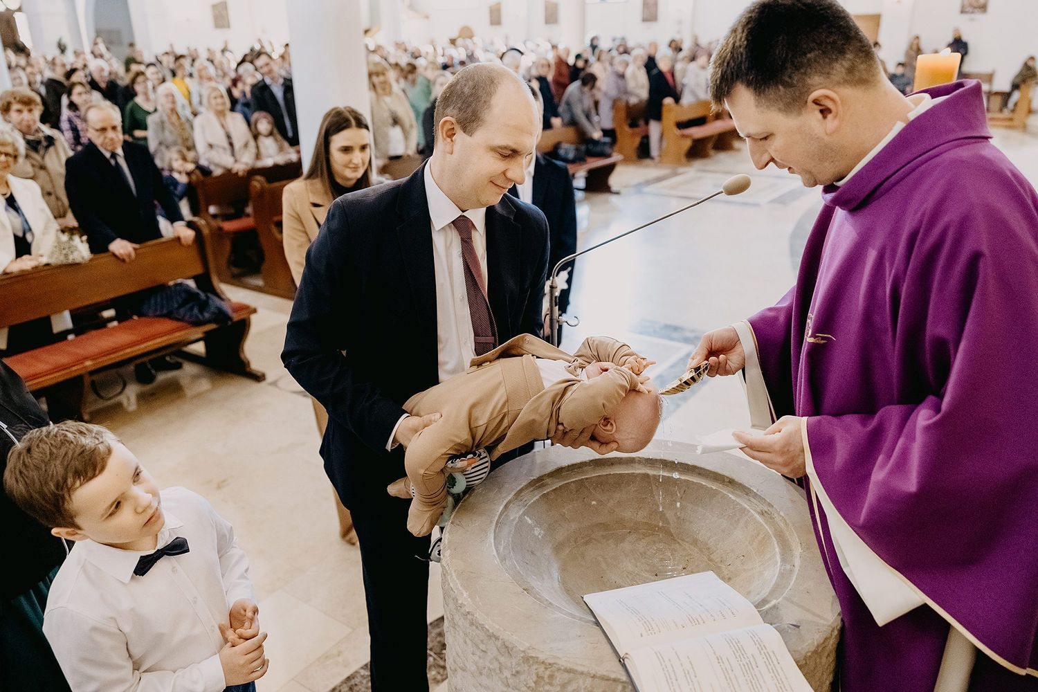 Catholic priest performing infant baptism at stone font with family and guests in church.