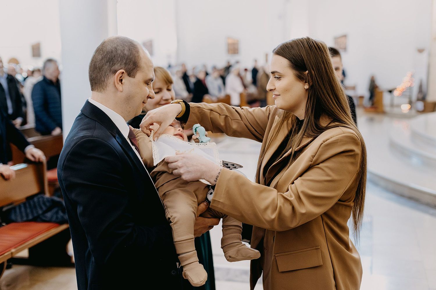 A couple in formal attire holds their baby during a church baptism ceremony.