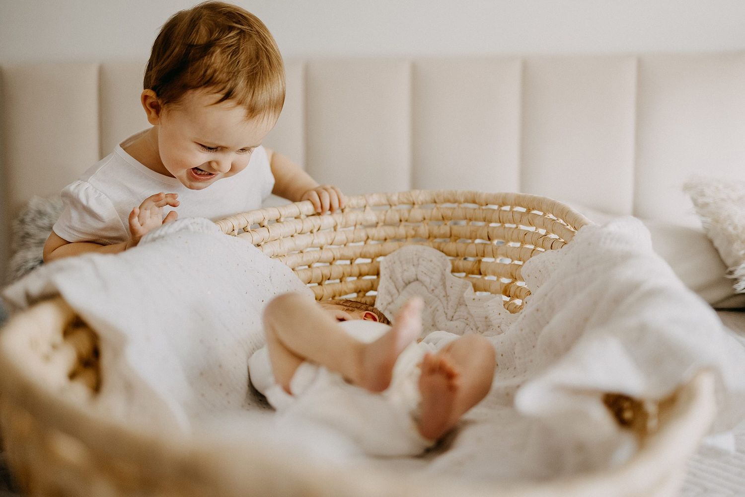 A curious toddler in white dress looks at a newborn baby resting in a wicker basket.