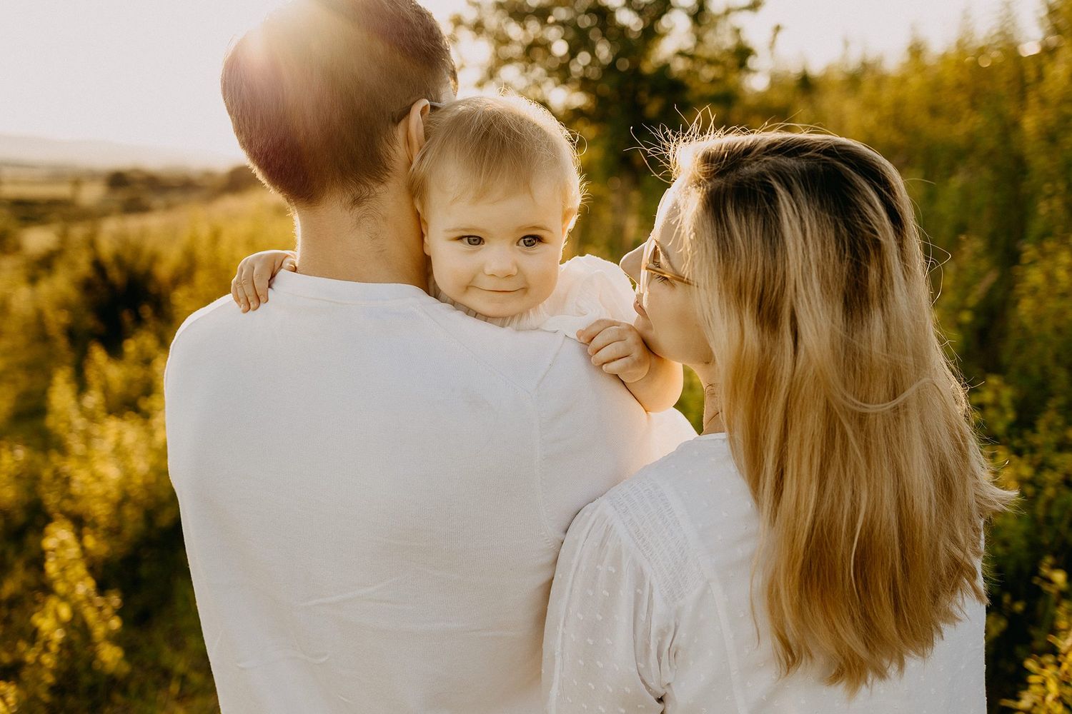 Mother kissing her toddler on the cheek while father holds the child in a sunny outdoor field.