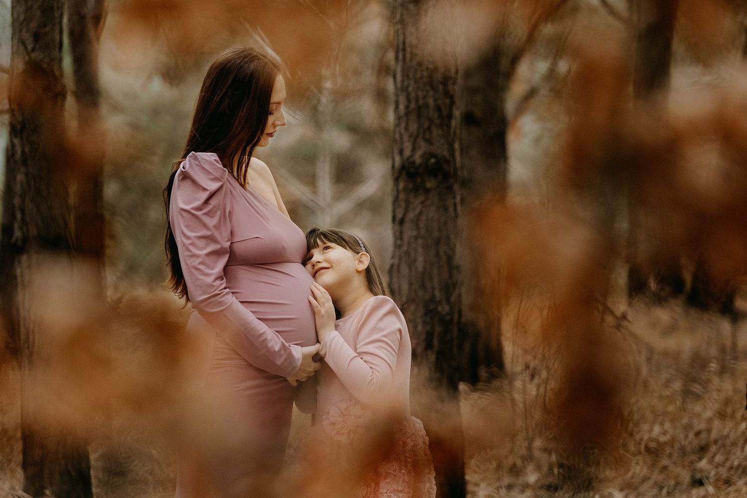 Young girl hugging pregnant mothers belly in an autumn forest during a maternity photo shoot.